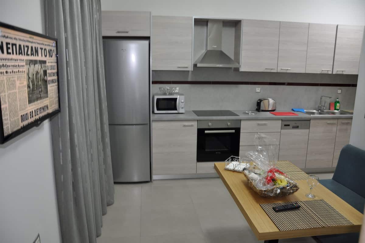 A modern kitchen with a refrigerator, microwave, oven, and sink. There is a table with a basket of groceries and a glass of wine.
