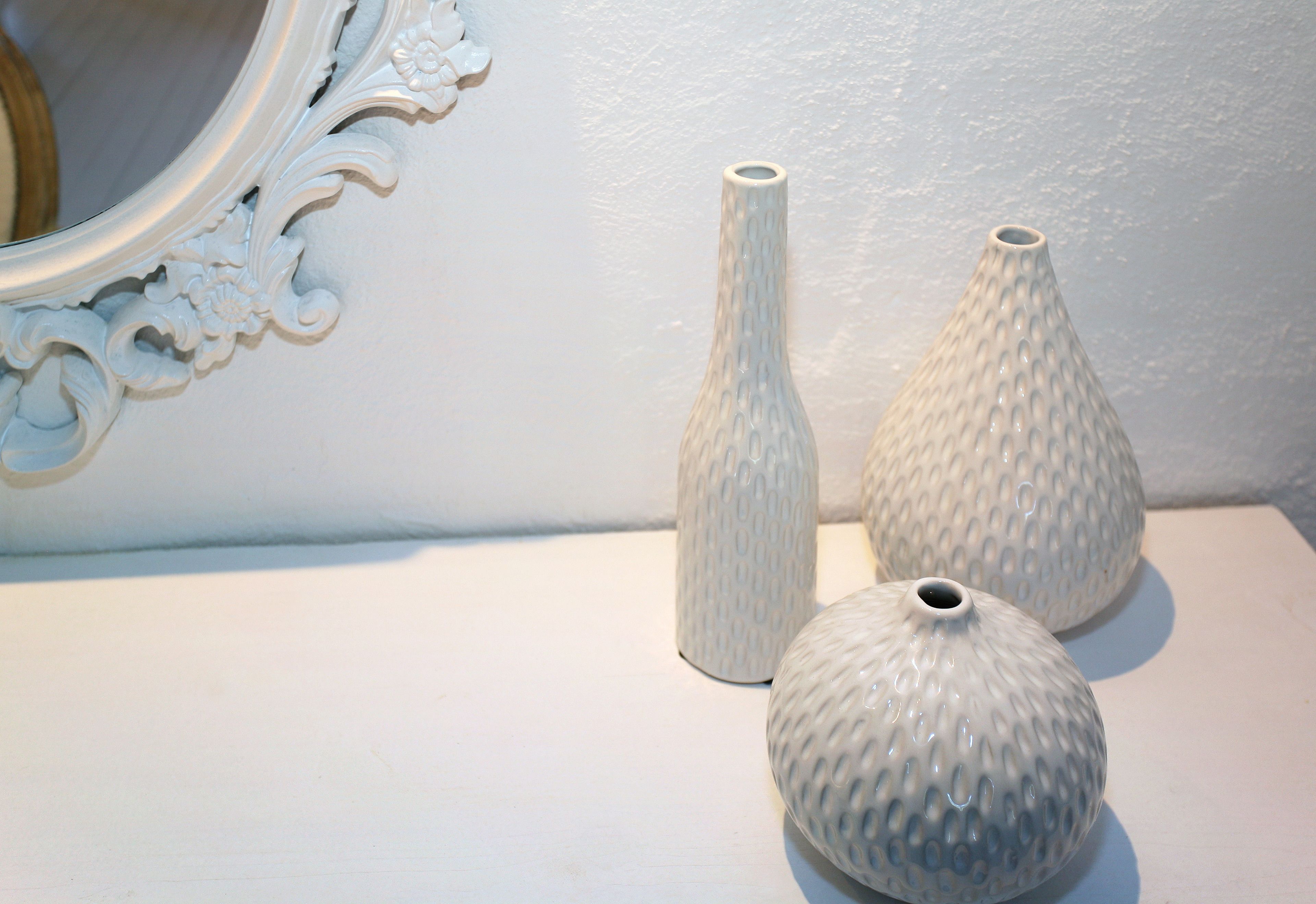 Three white ceramic vases on a white surface with a decorative mirror and wall in the background.