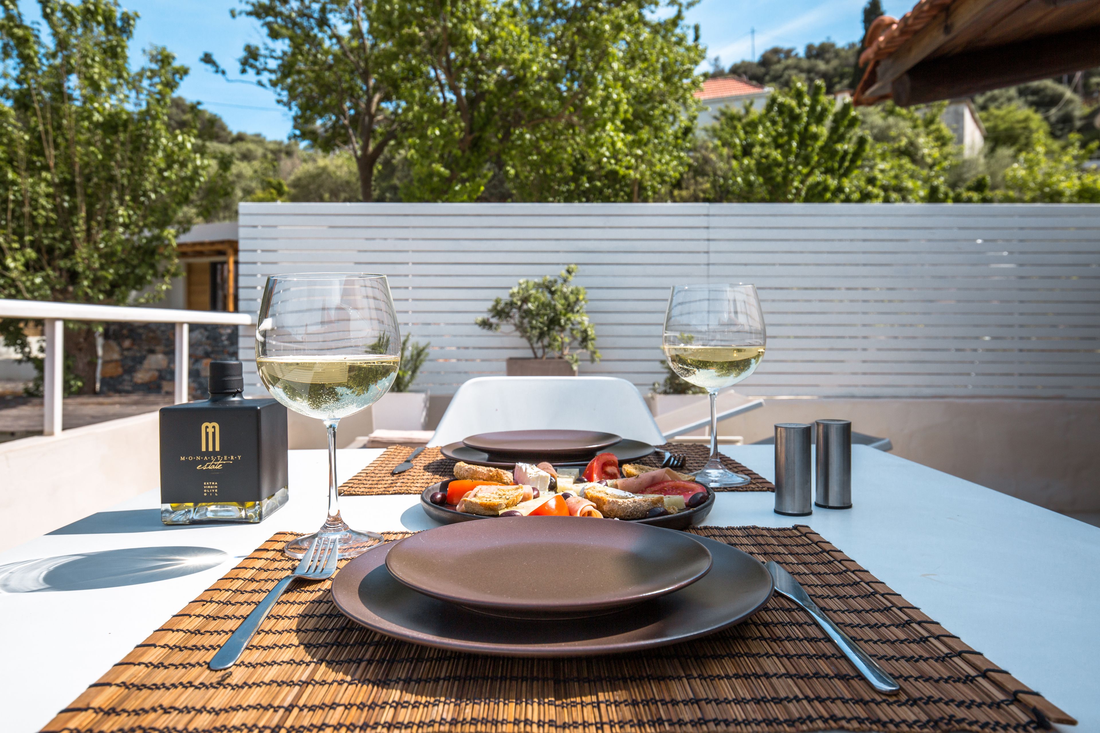 A table set with plates, cutlery, glasses, and a bottle of alcohol, with a view of trees and a building in the background.
