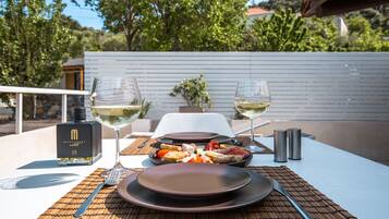 A table set with plates, cutlery, glasses, and a bottle of alcohol, with a view of trees and a building in the background.