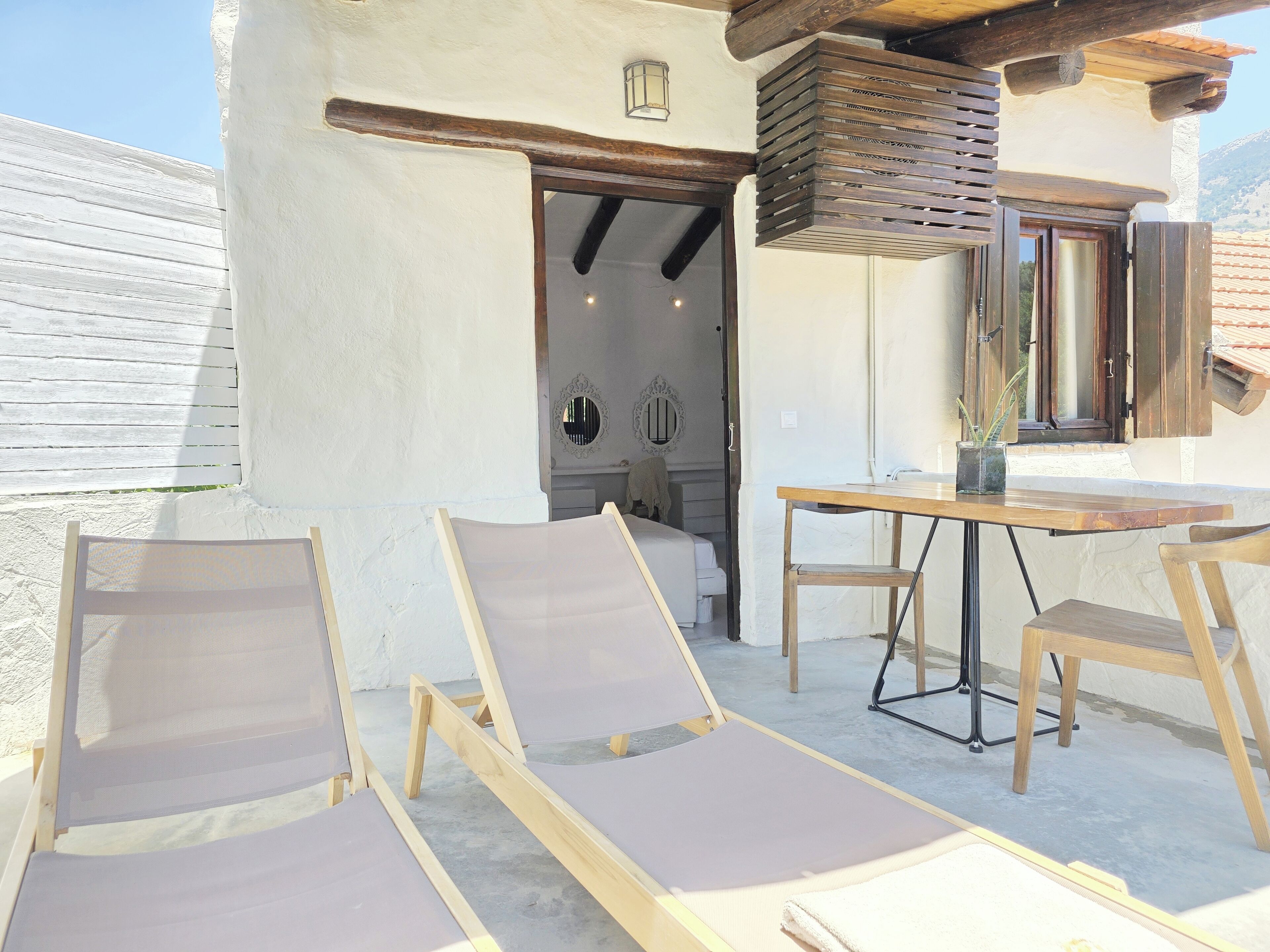 Un área de estar al aire libre iluminada por el sol, con sillones de madera y una mesa, una estructura de techo de madera y vistas del interior a través de un acceso abierto.