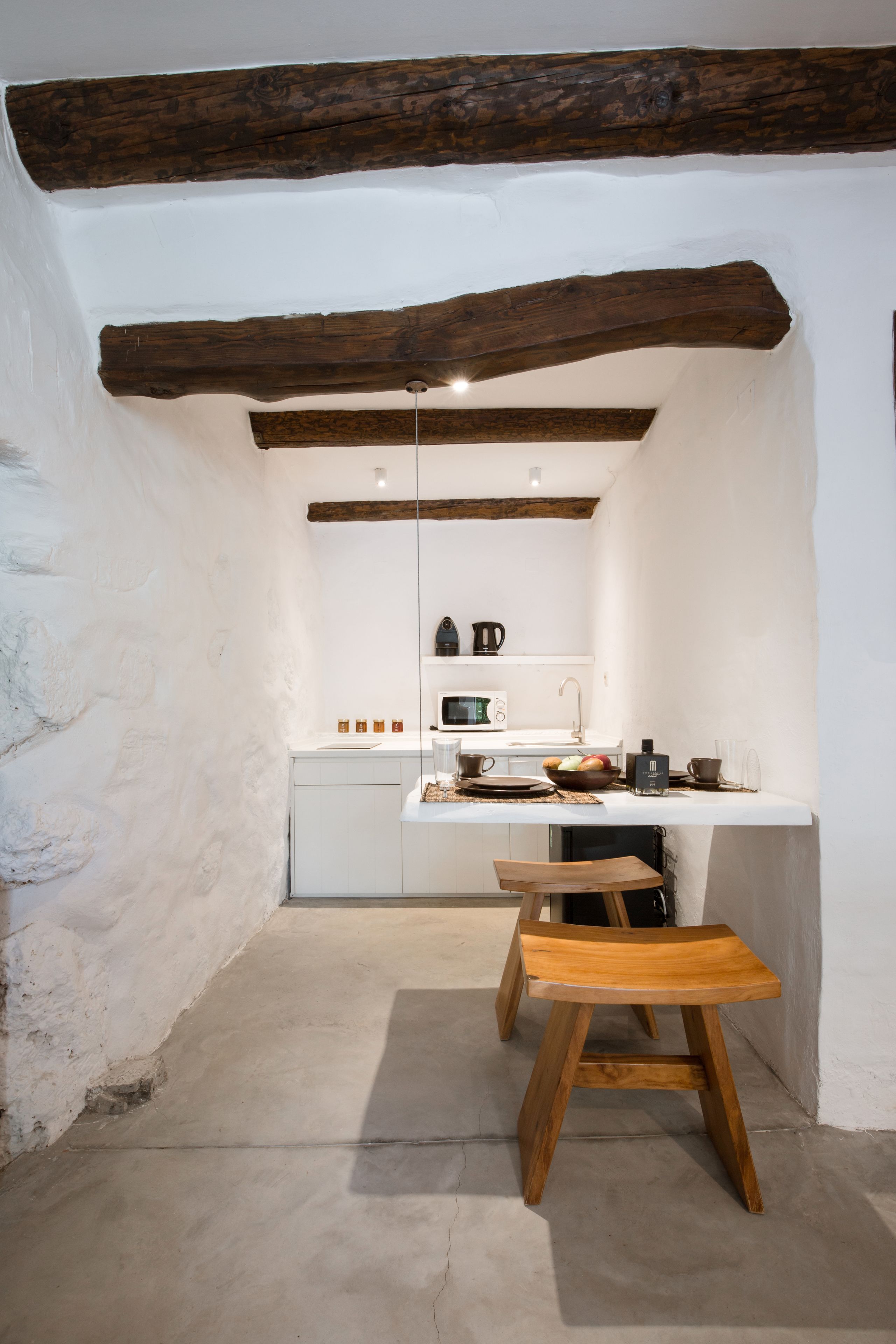 Una cocina compacta con paredes blancas, vigas de madera a la vista, una encimera blanca y un taburete de madera.