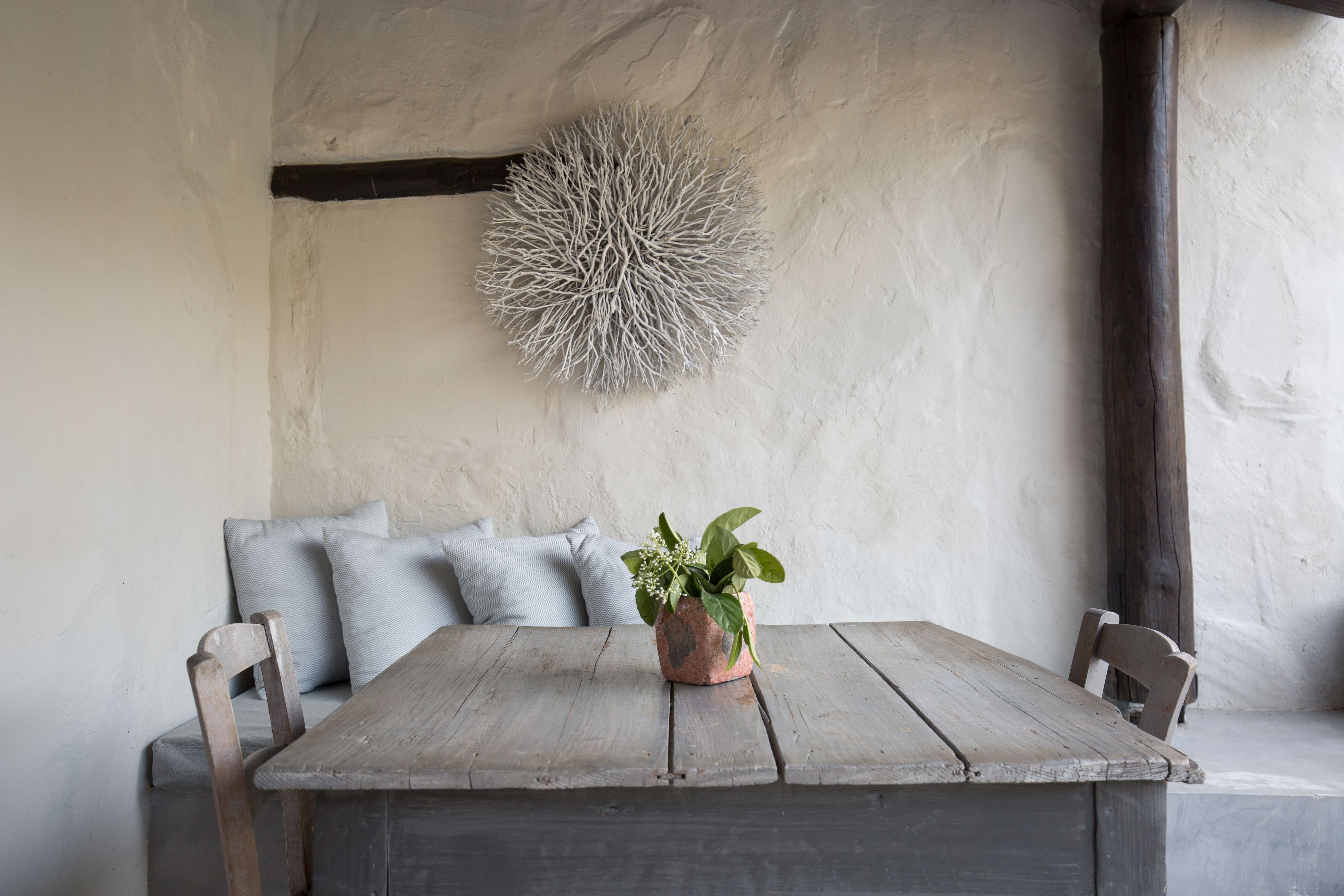 Una mesa de madera rústica con una planta en maceta, un banco acolchado con cojines y una pared texturizada con un adorno esférico.