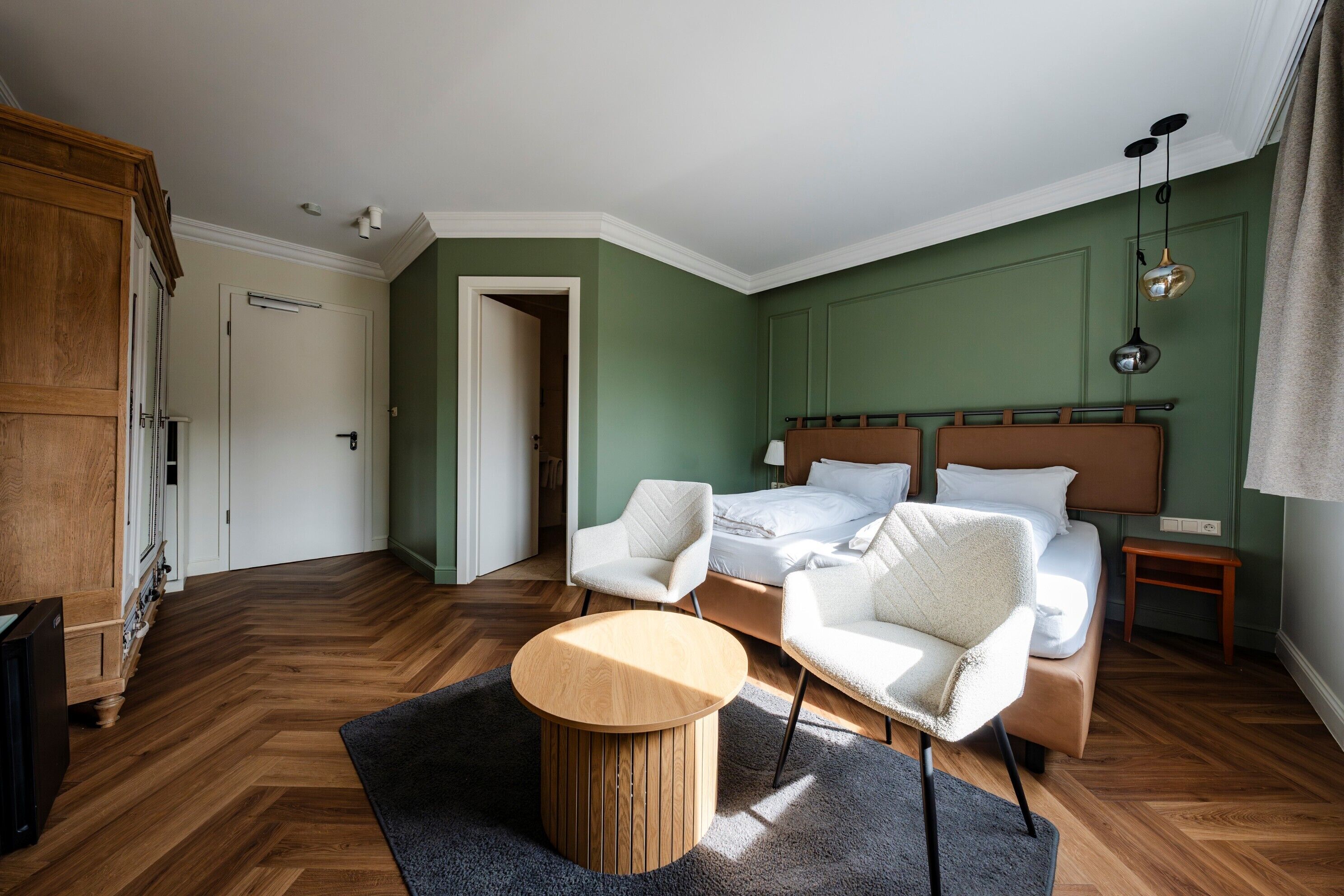 Une chambre à coucher comprenant un lit, deux fauteuils blancs, une table ronde en bois, une table de chevet et une armoire.