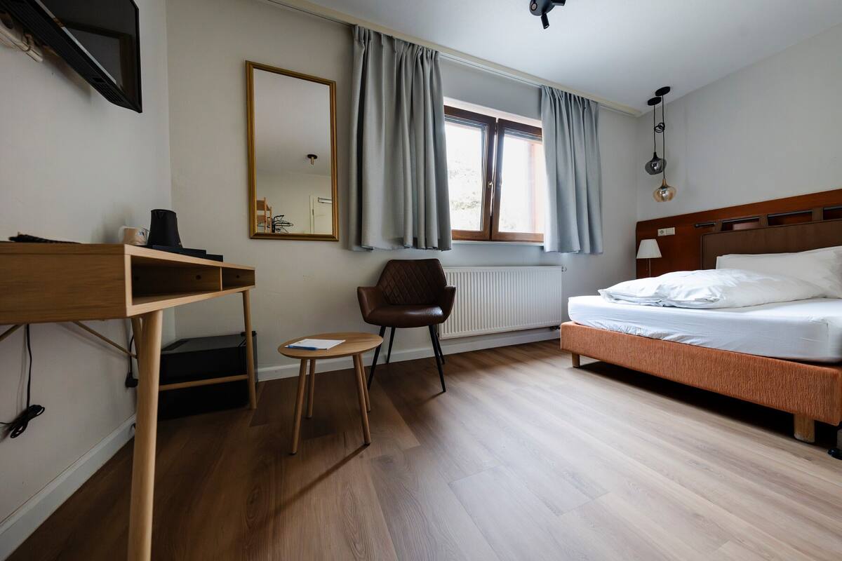 Ein Hotelzimmer mit einem Bett, einem Schreibtisch mit Stuhl, einem Heizkörper, einem Fenster mit Vorhängen und einem Spiegel an der Wand.