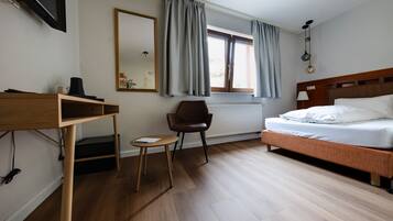 A hotel room with a bed, a desk with a chair, a radiator, a window with curtains, and a mirror on the wall.