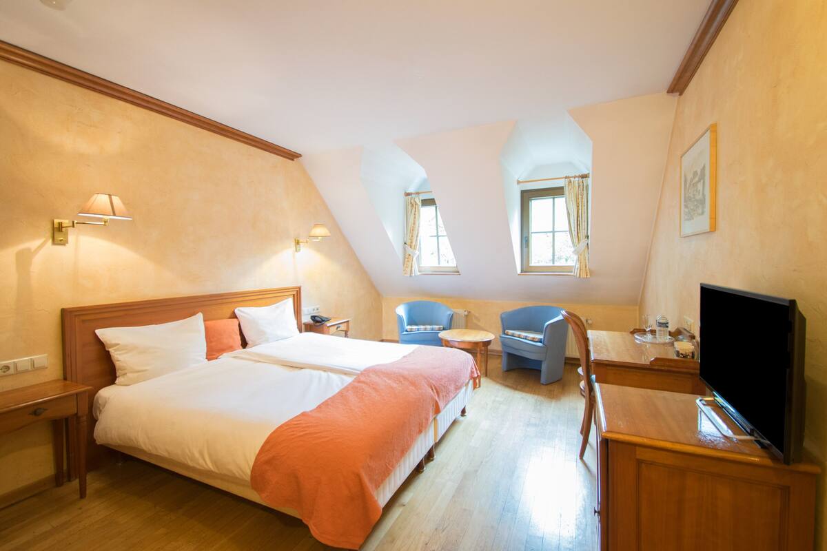 Ein Hotelzimmer mit einem großen Bett, einem Schreibtisch, einem Sessel, einem Fernseher und zwei Fenstern.