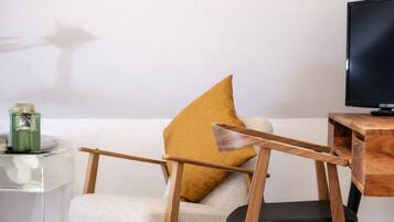 A modern living room with a minimalist design, featuring a wooden chair with a unique backrest, a mustard-colored pillow, a glass coffee table, and a flat-screen TV on a wooden stand.