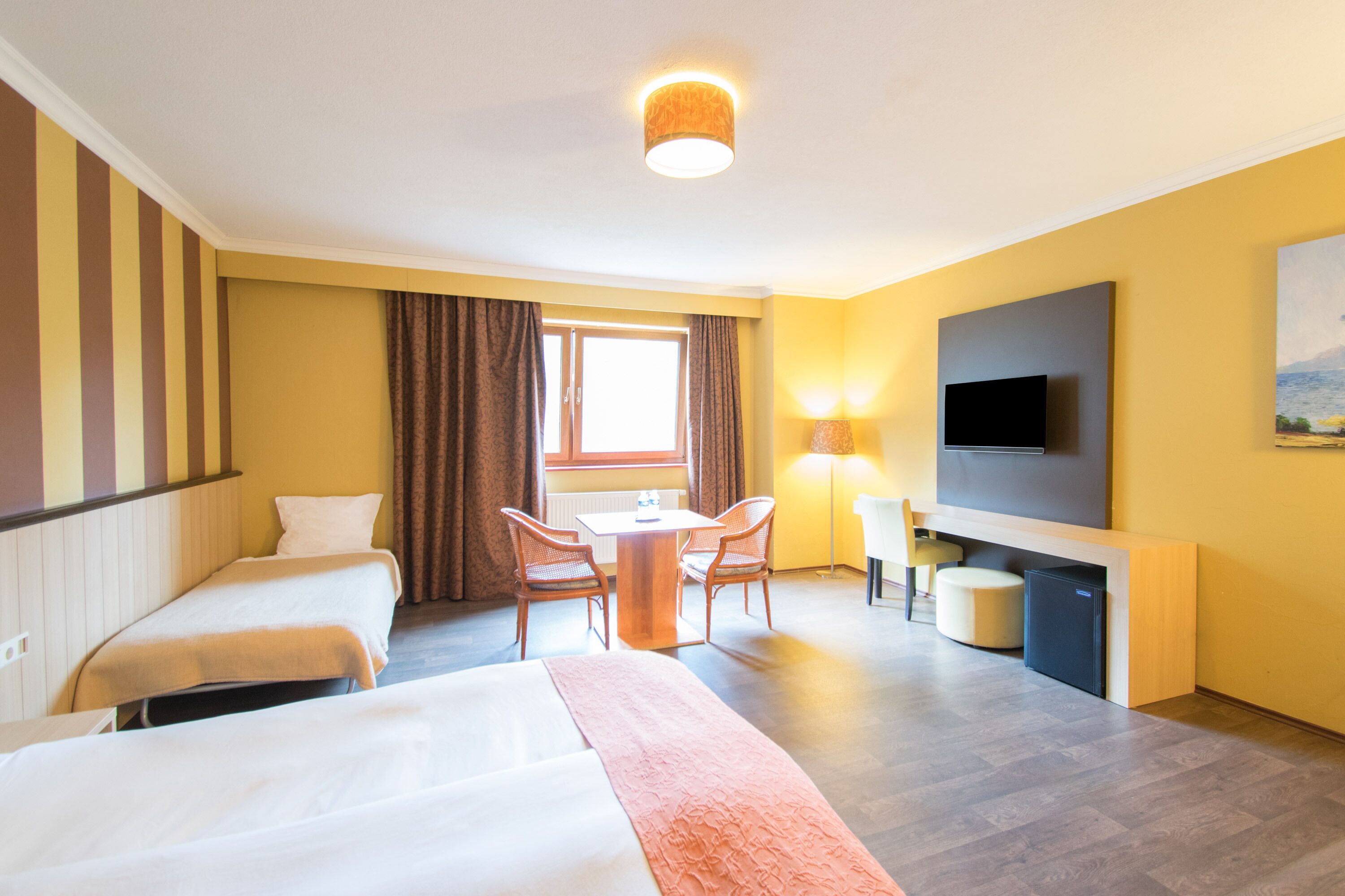 A hotel room with two beds, a desk, chairs, a TV, and a window with curtains.