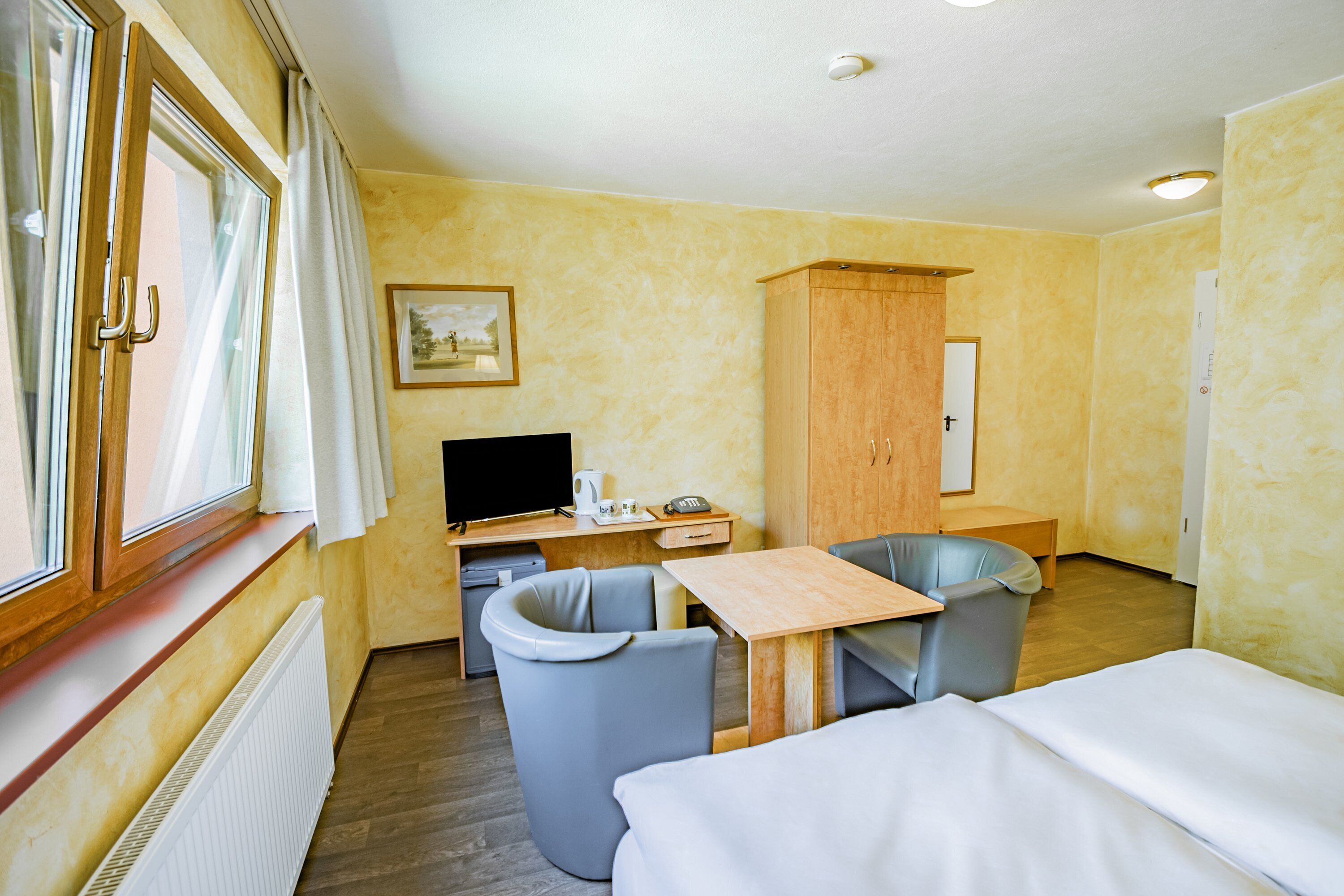 Hotellihuone, jossa on sänky, työpöytä tietokoneella, tuoli, vaatekaappi ja ikkuna, jossa on verhot.