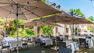 An outdoor dining area with tables and chairs under large umbrellas, surrounded by greenery and a building with a classic architectural style.