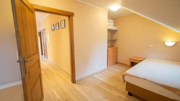 Quarto com piso de madeira, cama com cobertura branca, mesa de cabeceira e luminária fixada na parede.