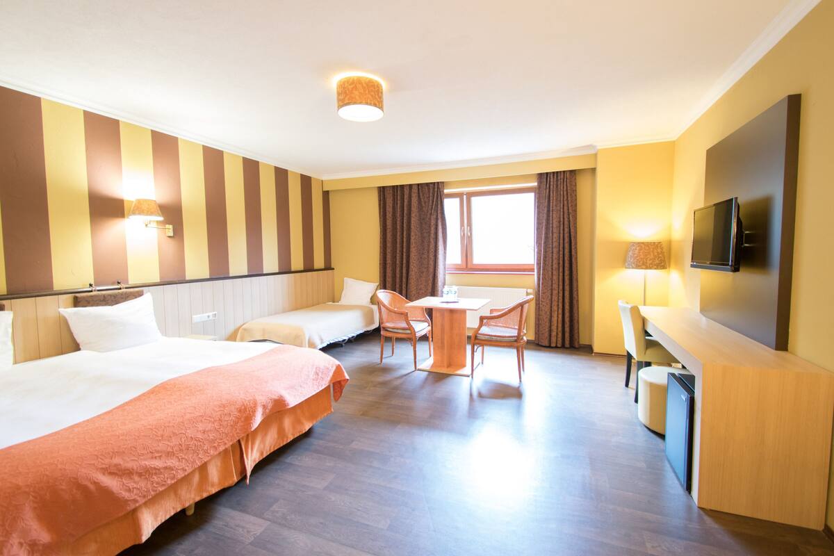 Ein Hotelzimmer mit zwei Betten, einem Esstisch mit Stühlen, einem Fernseher und einem Fenster mit Vorhängen.