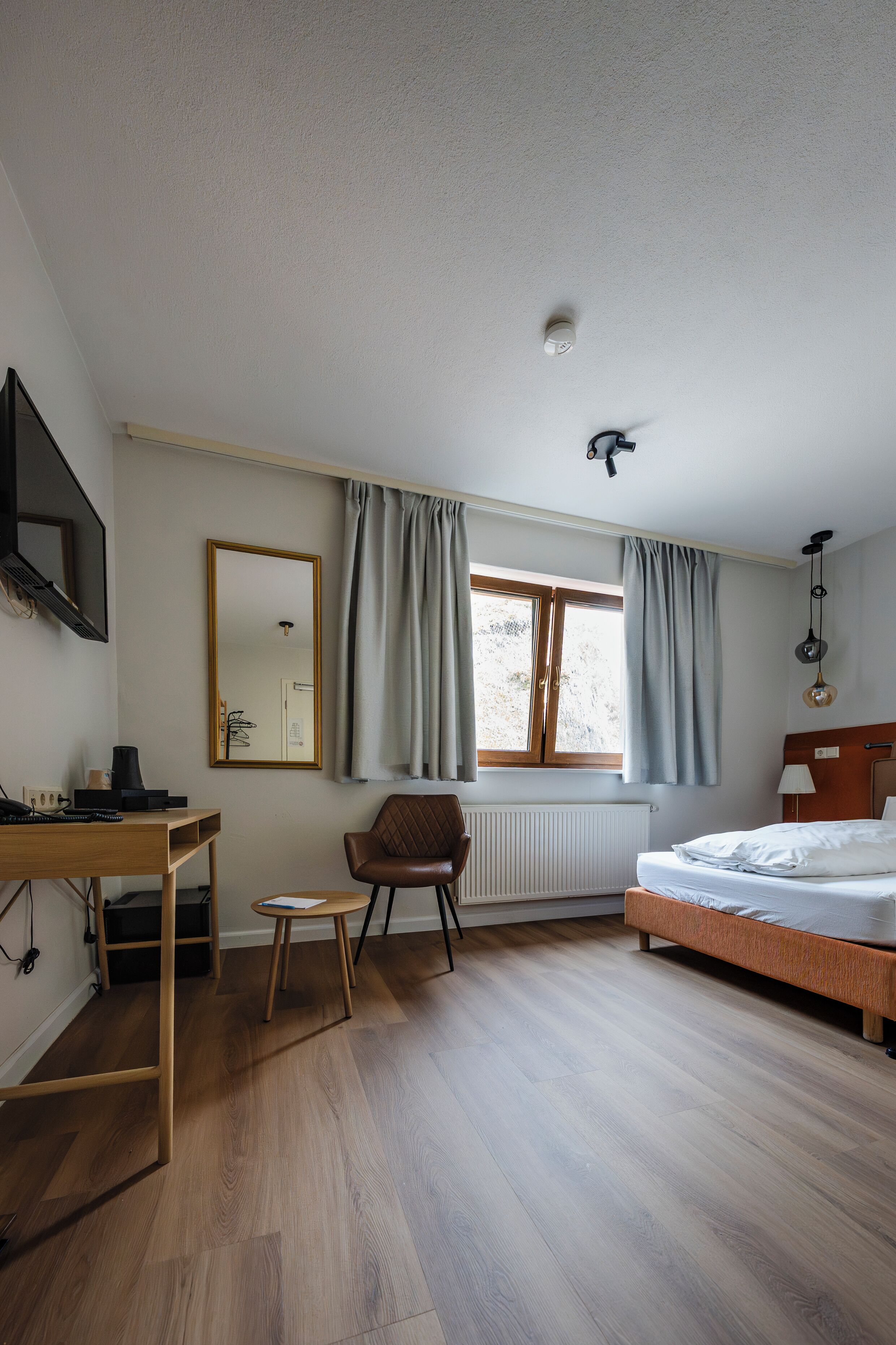 A bedroom with a bed, a desk, a chair, a window with curtains, a radiator, and a ceiling light fixture.