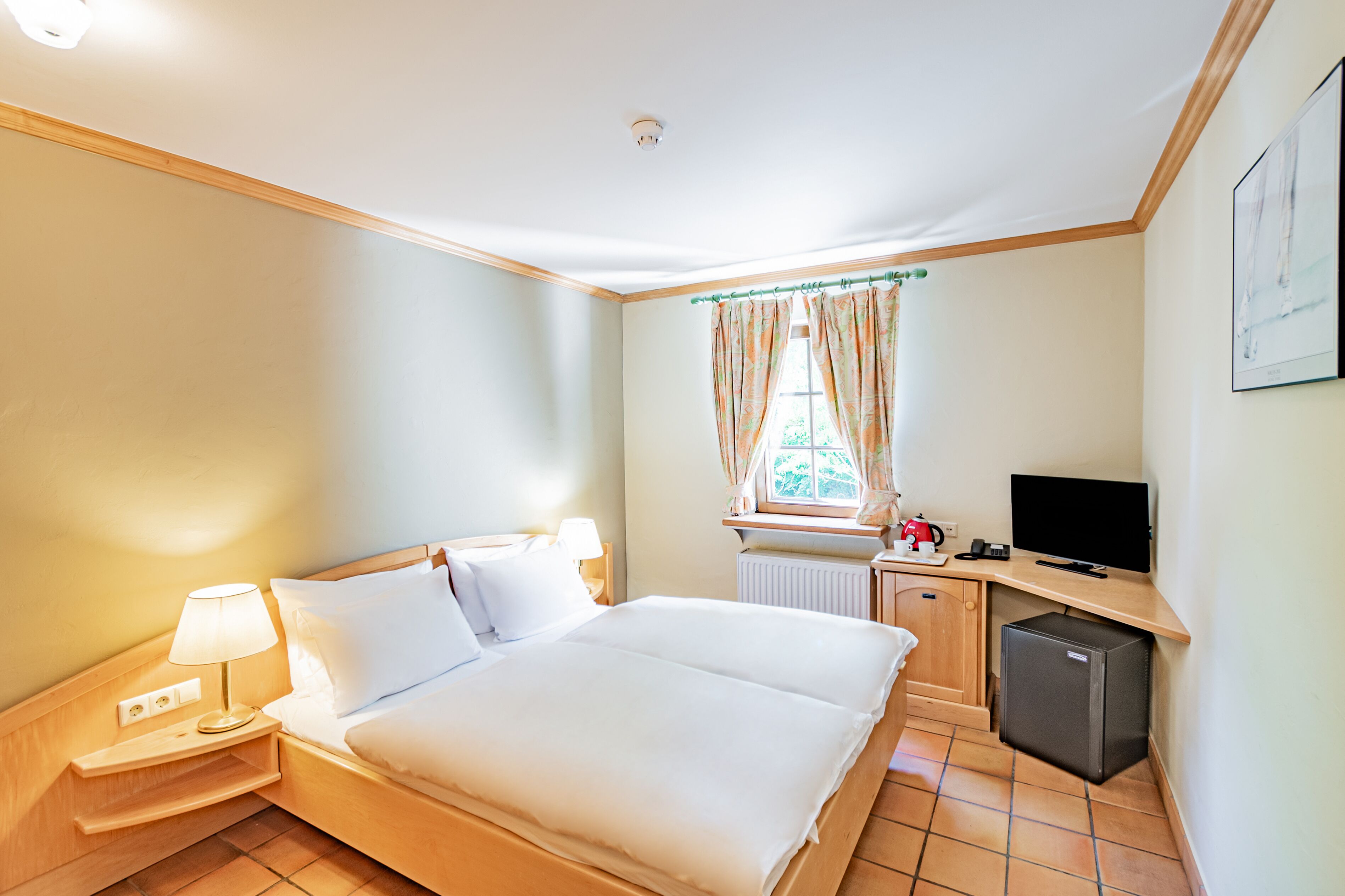 A hotel room with a bed, bedside lamp, a small refrigerator, a desk with a television, and a window with curtains.