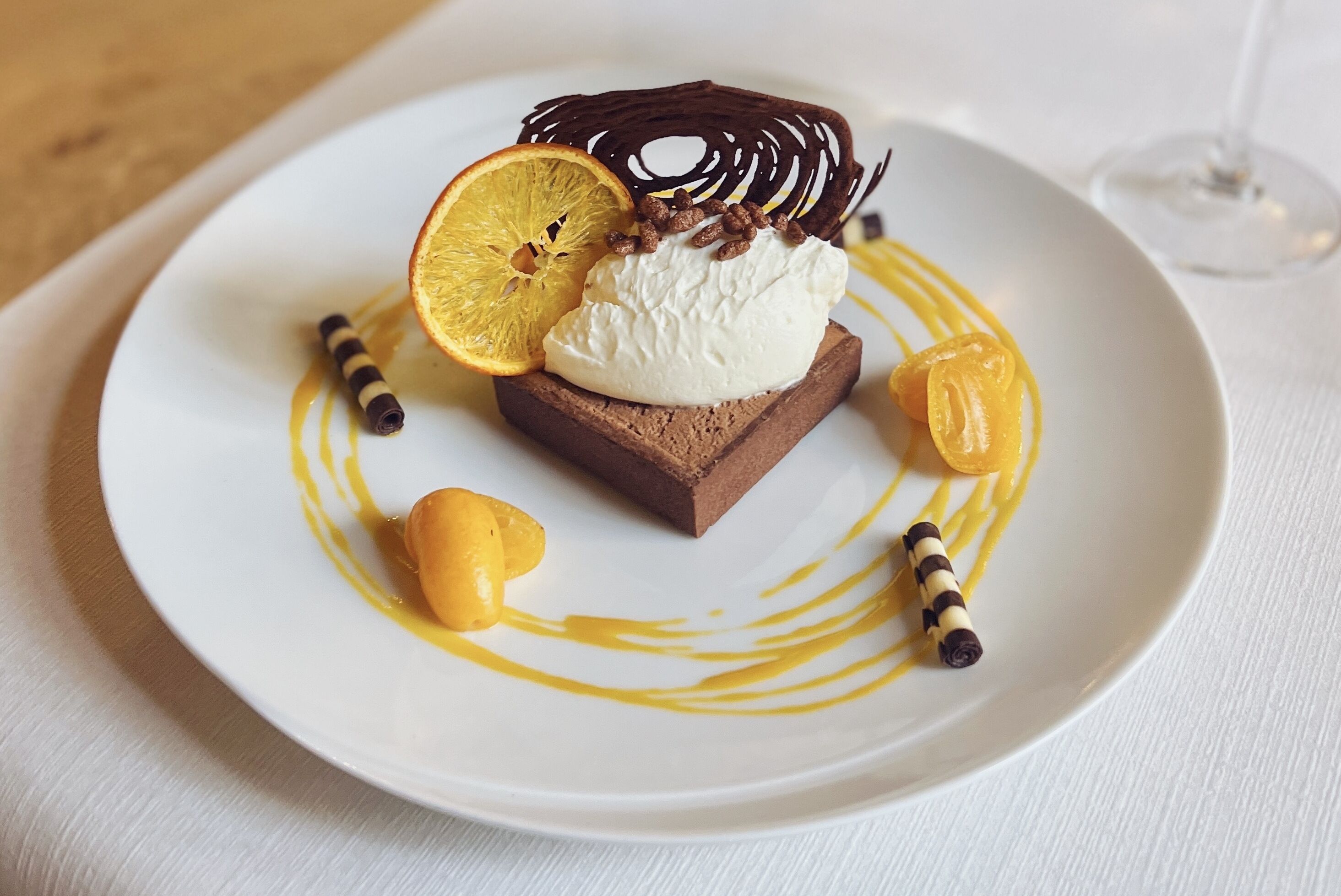 A dessert on a white plate with a chocolate base, white cream, orange slice, and chocolate drizzle.