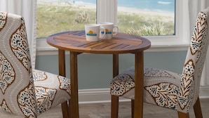 A round wooden table with two patterned chairs, a window with a view of the beach, and two white mugs on the table.