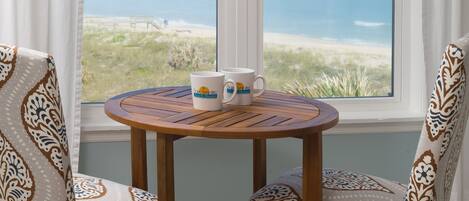 Una mesa redonda de madera con dos sillas estampadas, una ventana que da a la playa y dos tazas blancas sobre la mesa.