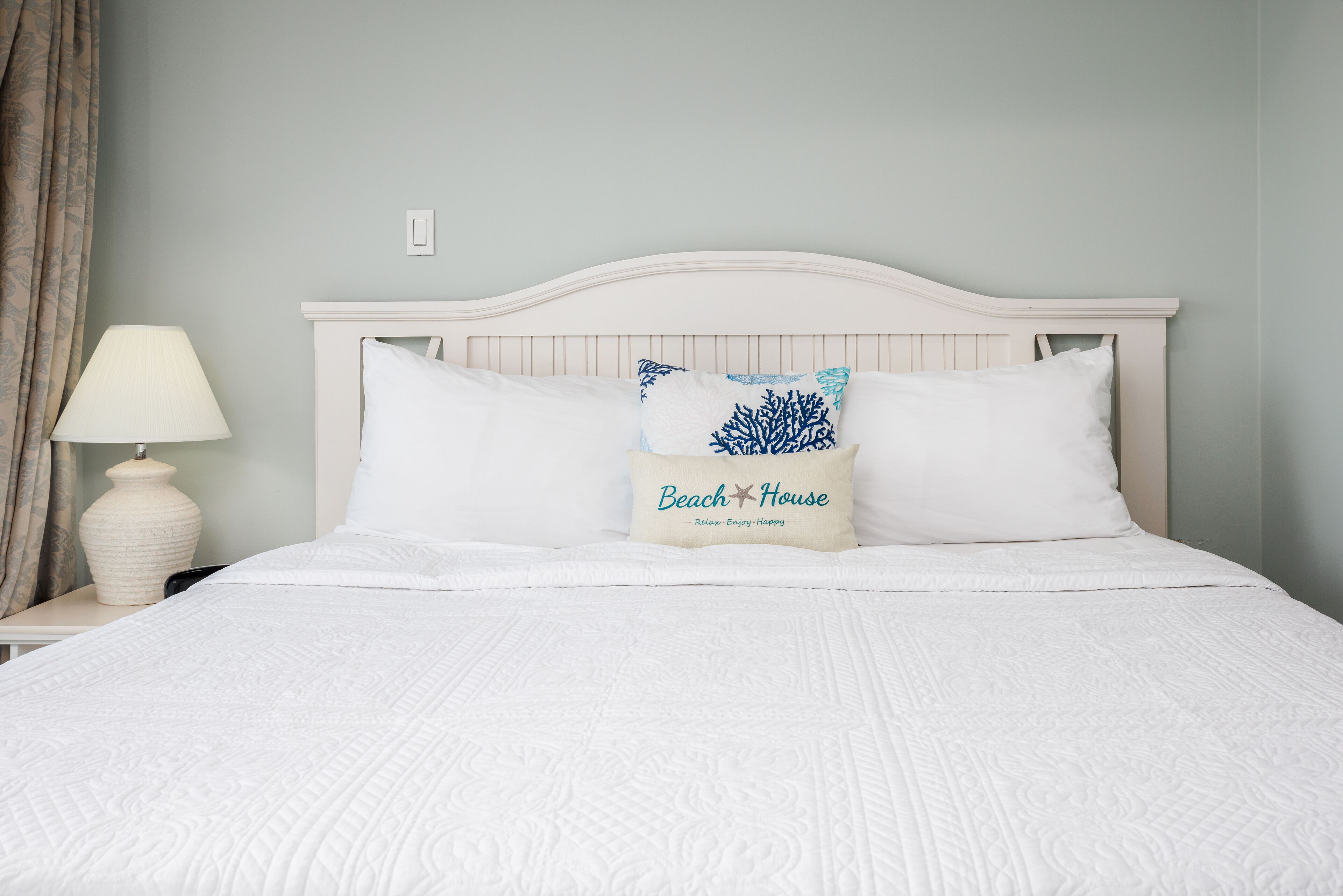 Una cama bien hecha con ropa blanca y dos almohadones decorativos, uno con motivos playeros, en una habitación con paredes verde claro y cabecera blanca.