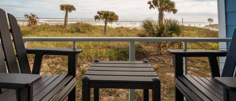 A balcony with two wooden chairs and a small table, overlooking a beach with palm trees and the ocean.