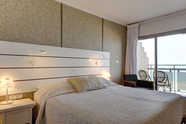 Habitación de hotel con una cama grande, una mesita de noche con lámpara, una silla y vista al mar desde un balcón.