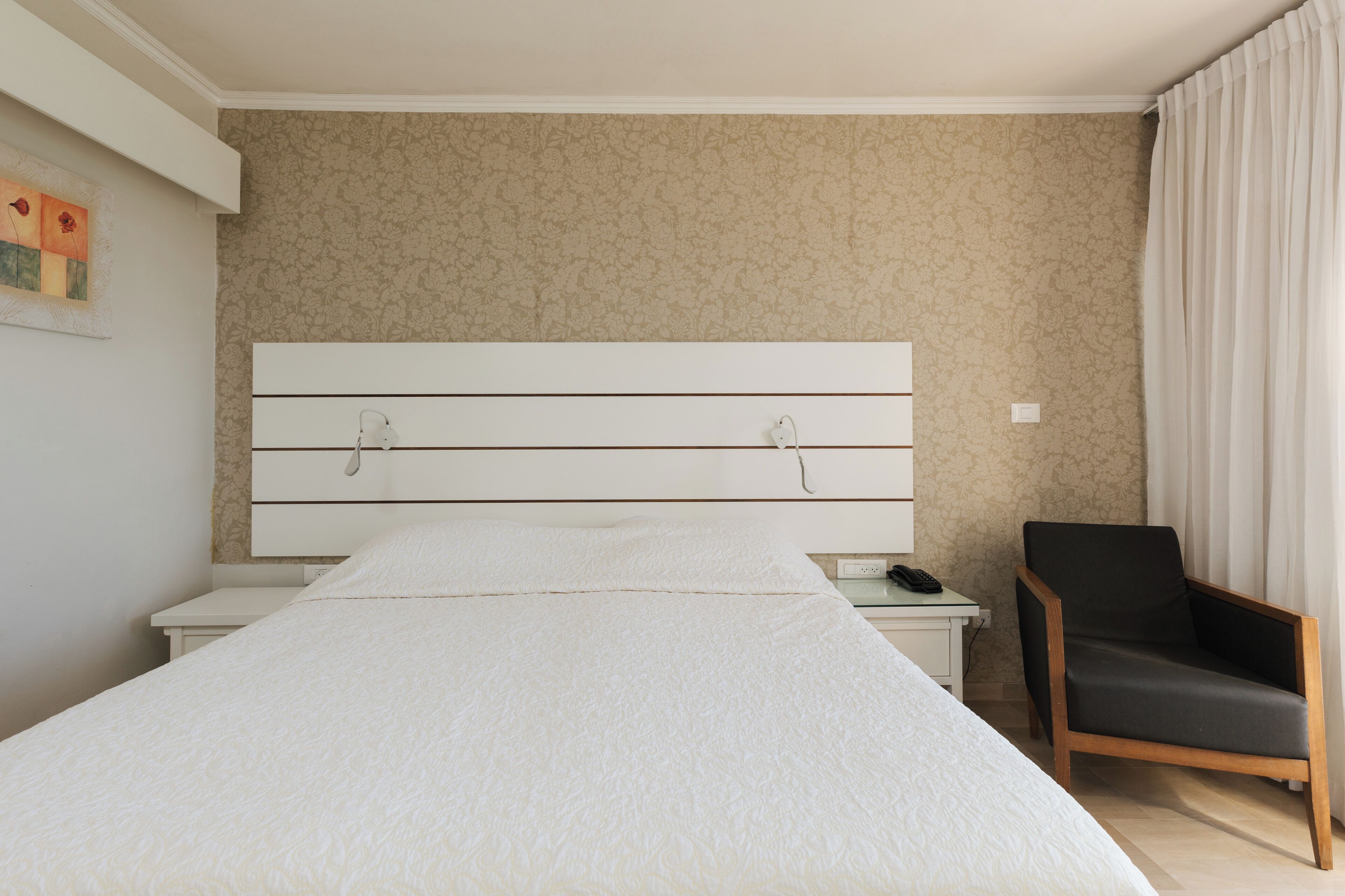 A hotel room with a large bed, a nightstand, a chair, and a wall with a floral pattern.