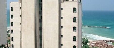 Un edificio grande, de color beige, con un elemento vertical central y múltiples ventanas, situado cerca de una playa con vista despejada al océano.