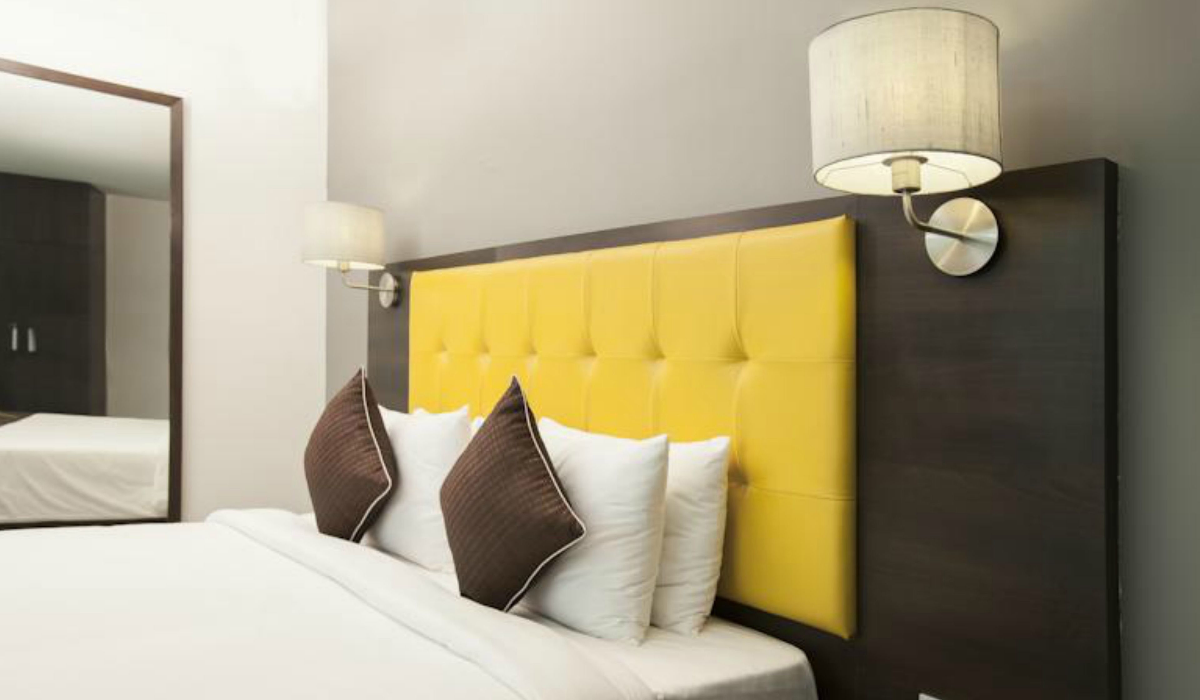 Une chambre d’hôtel avec une tête de lit jaune à motifs capitonnés, du linge de lit blanc et une lampe de chevet.