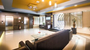 Un hall de hotel con mostrador de recepción, zona de estar, ascensores y un gran ventanal con persianas.