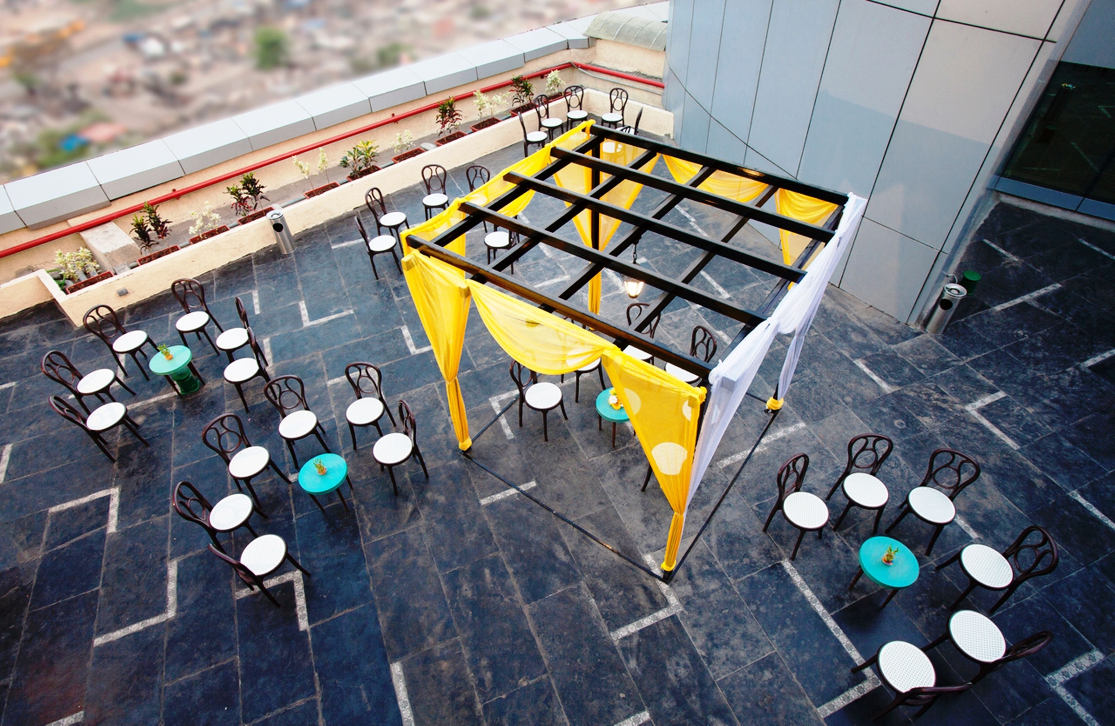 A rooftop terrace with tables and chairs arranged for dining, surrounded by yellow drapes and potted plants.