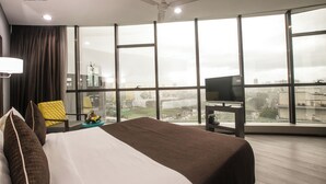 Habitación de hotel con una cama grande, televisión, ventilador de techo y vistas de la ciudad a través de amplios ventanales.