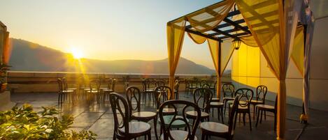 A rooftop terrace with tables and chairs, a pergola with draped fabric, and a view of the sun setting over the mountains.