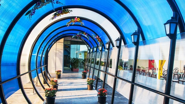 Un paseo techado con techo azul traslúcido, plantas en macetas y zonas de estar al aire libre.