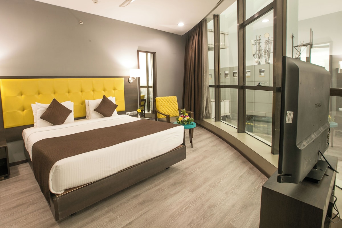 A hotel room with a large bed, a desk, a chair, a TV, and a window with a city view.