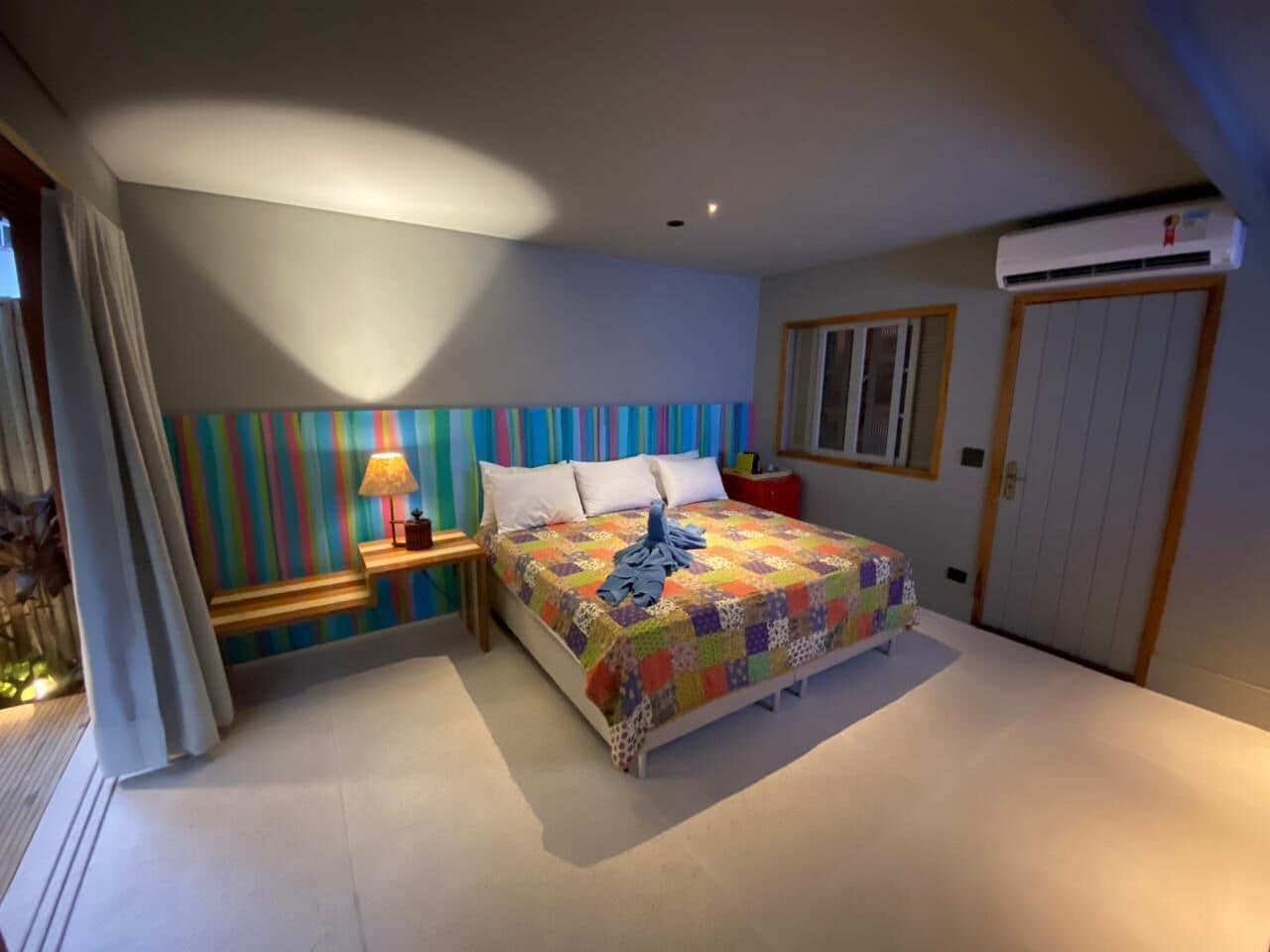 Un dormitorio con una cama, una mesita de noche con lámpara, una pared de rayas coloridas y una puerta con unidad de aire acondicionado.