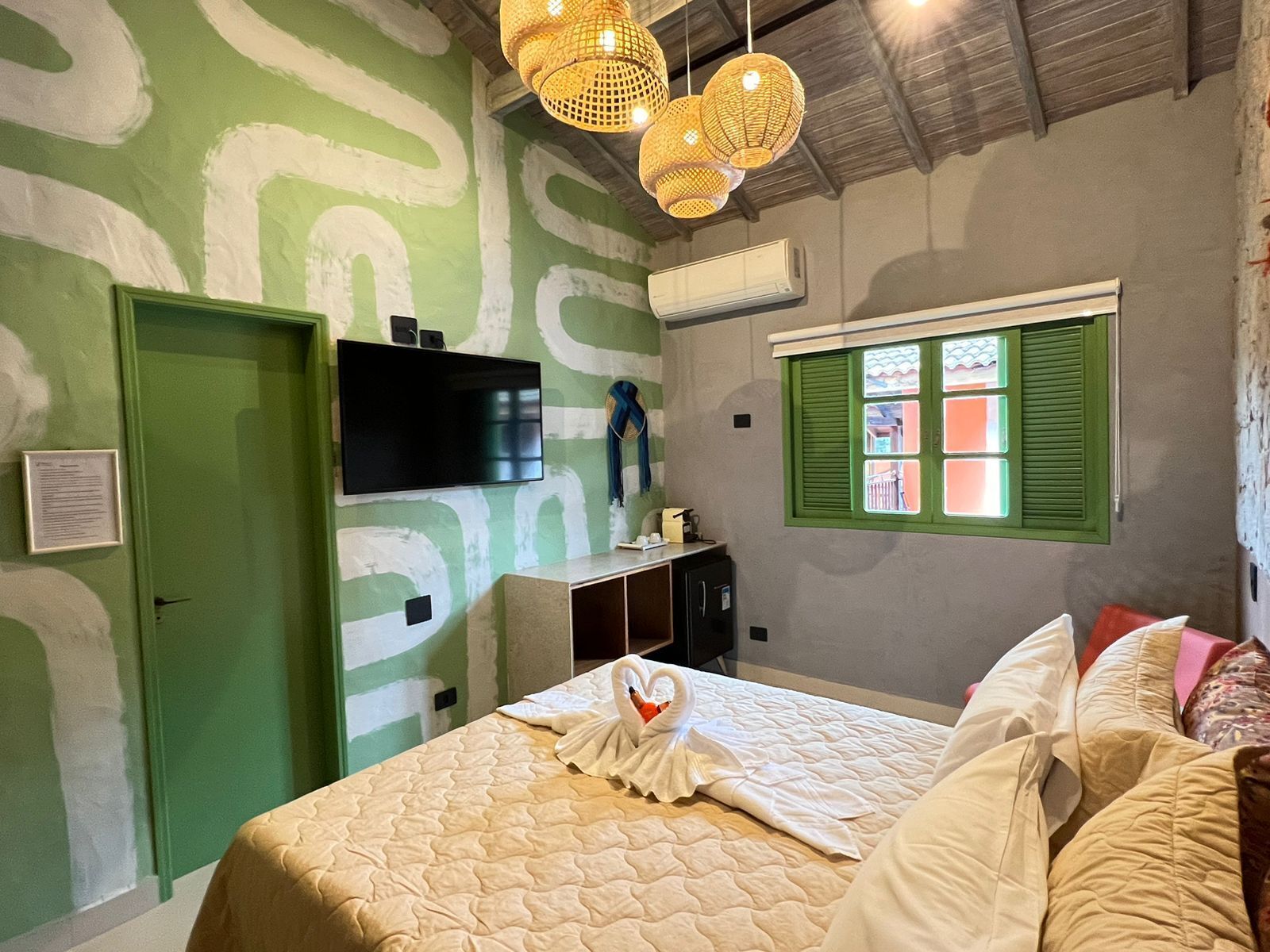 Een hotelkamer met een groene deur, een flatscreen televisie, een raam met groene luiken en een bed met een zwaan-vormig kussen.
