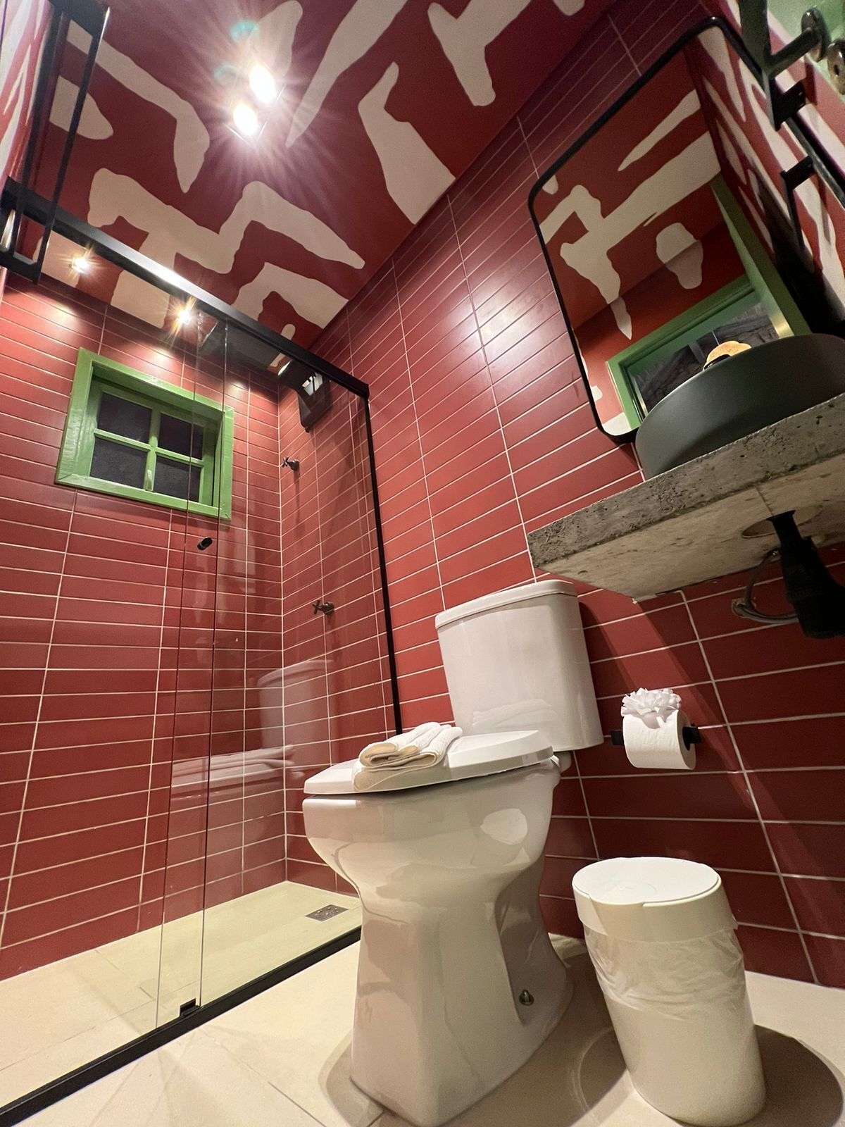 Une salle de bain avec des murs recouverts de carreaux rouges, des toilettes blanches, une fenêtre à cadre vert et un lavabo avec un miroir au-dessus.