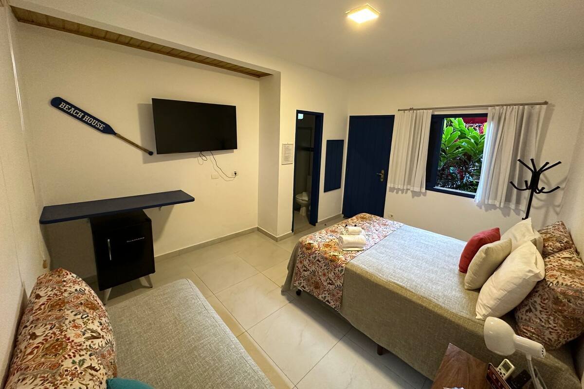 A hotel room with a TV, a desk, a bed with a canopy, a window with curtains, and a coat rack.