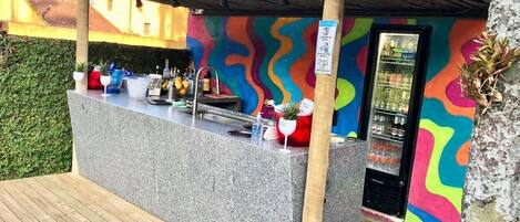 A tiki bar with a colorful mural, a counter with drinks, and a small refrigerator.