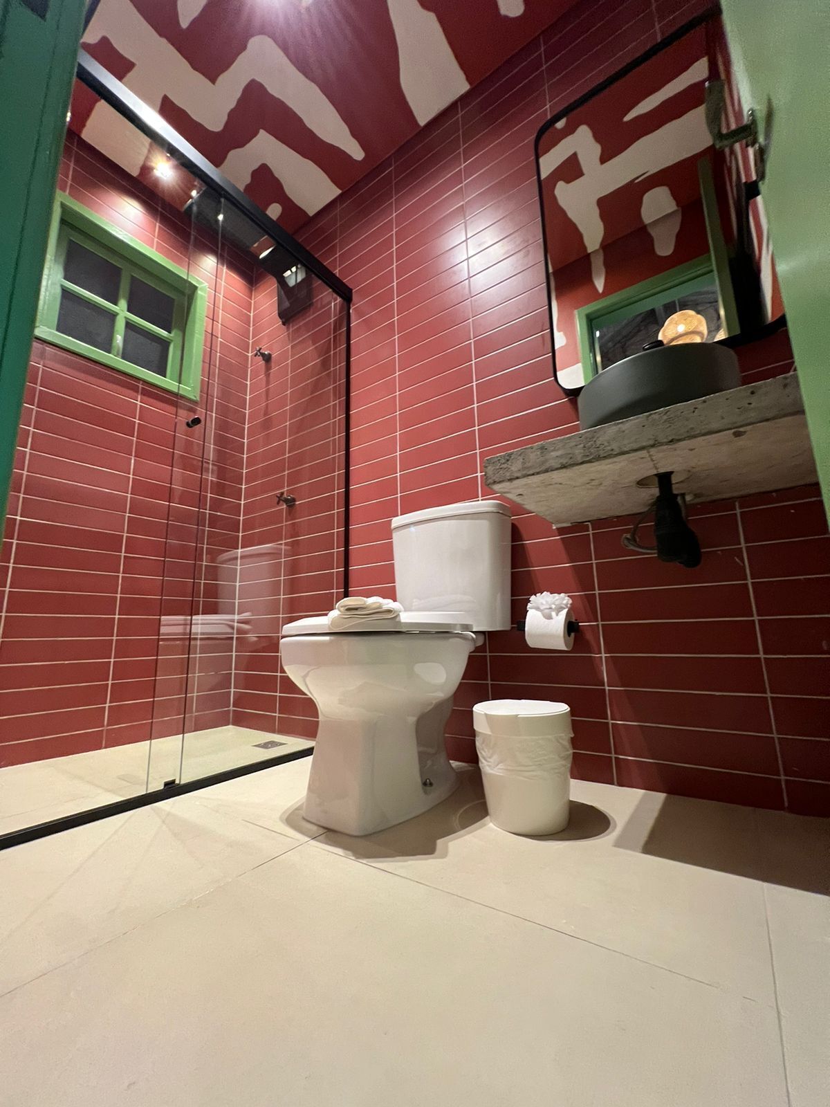 A bathroom with red tiled walls, a white toilet, a white bucket, and a shelf with a black pot.