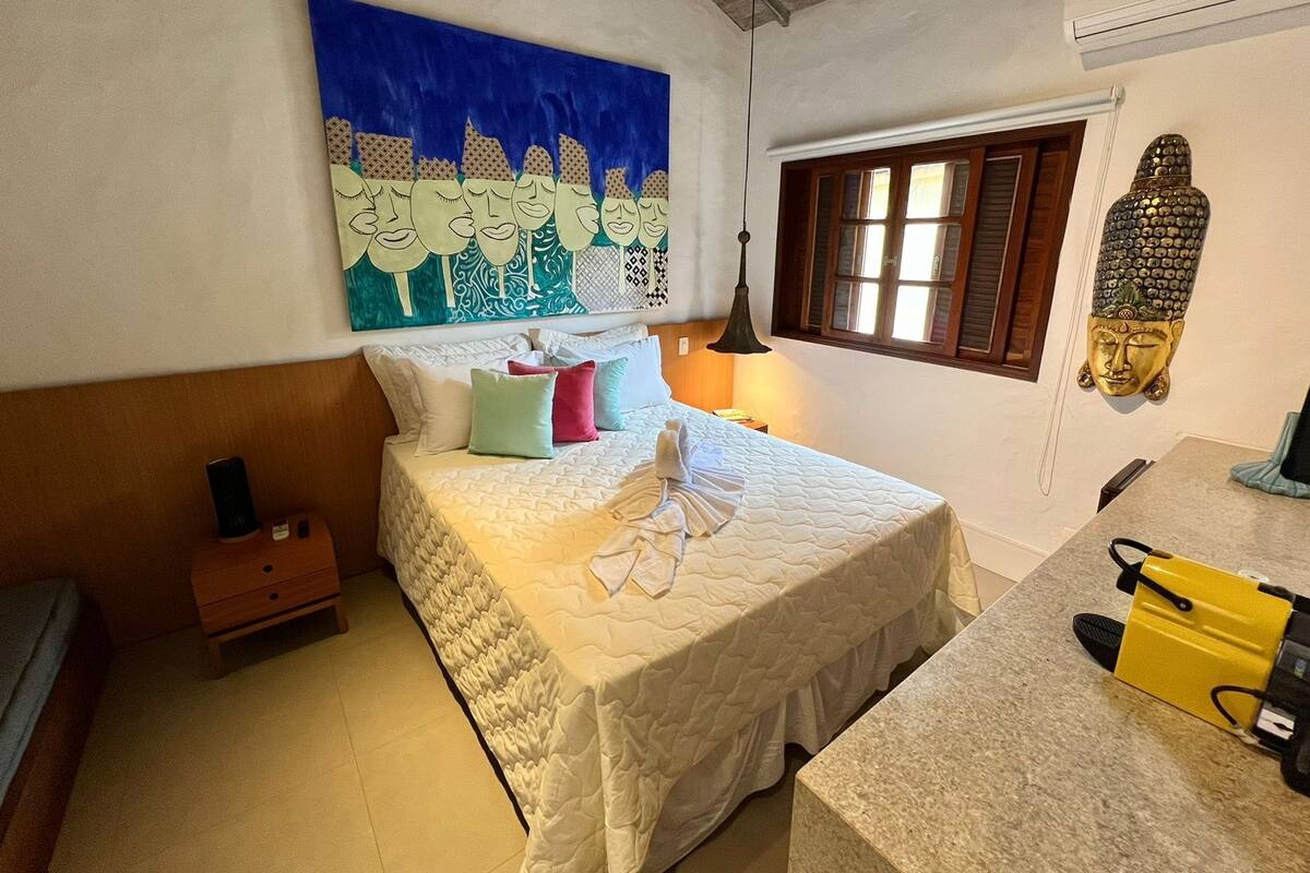 A bedroom with a bed, pillows, a nightstand, a lamp, a Buddha statue, and a window.