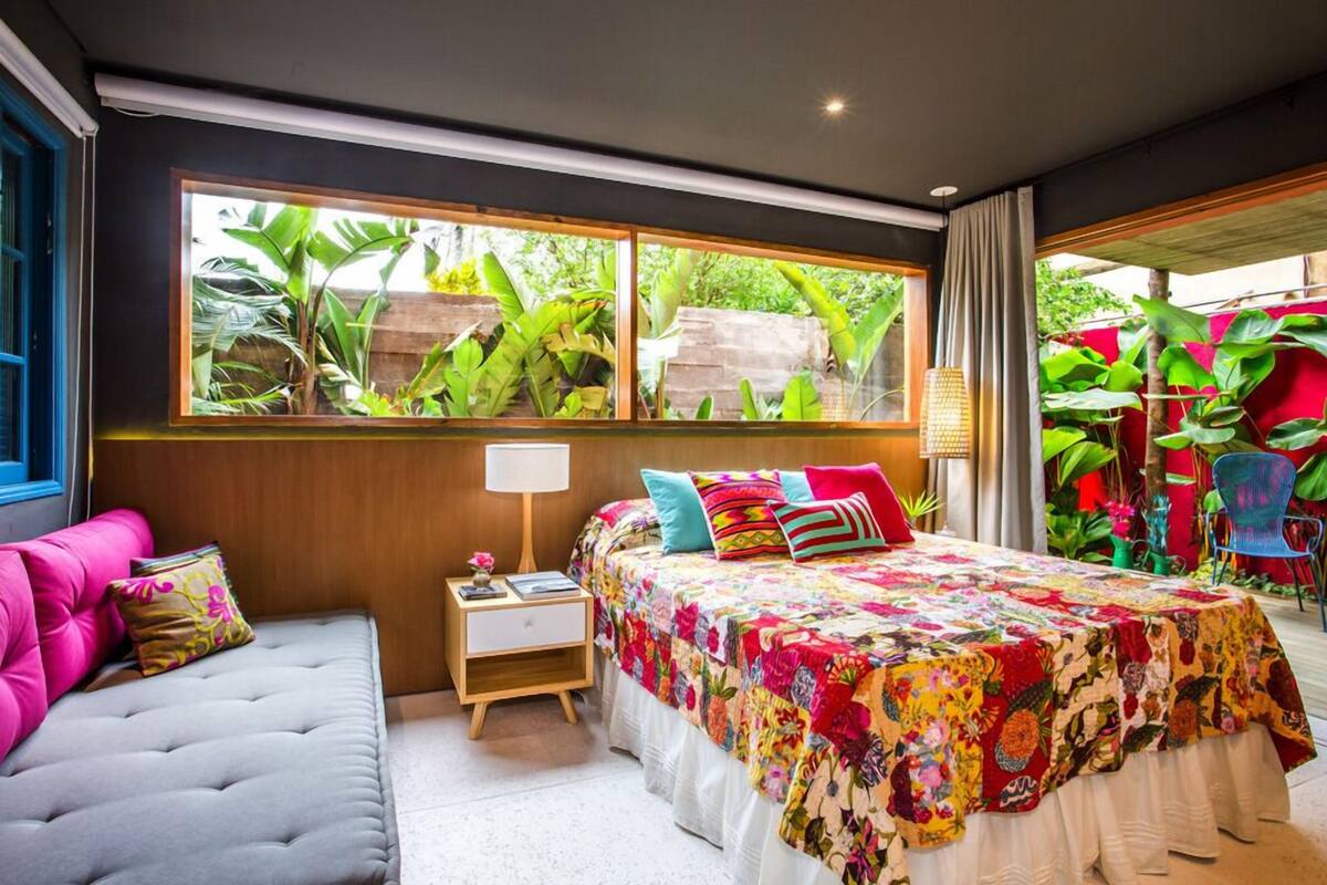 A bedroom with a large window, a bed with colorful bedding, a grey sofa, and a small bedside table with a lamp.