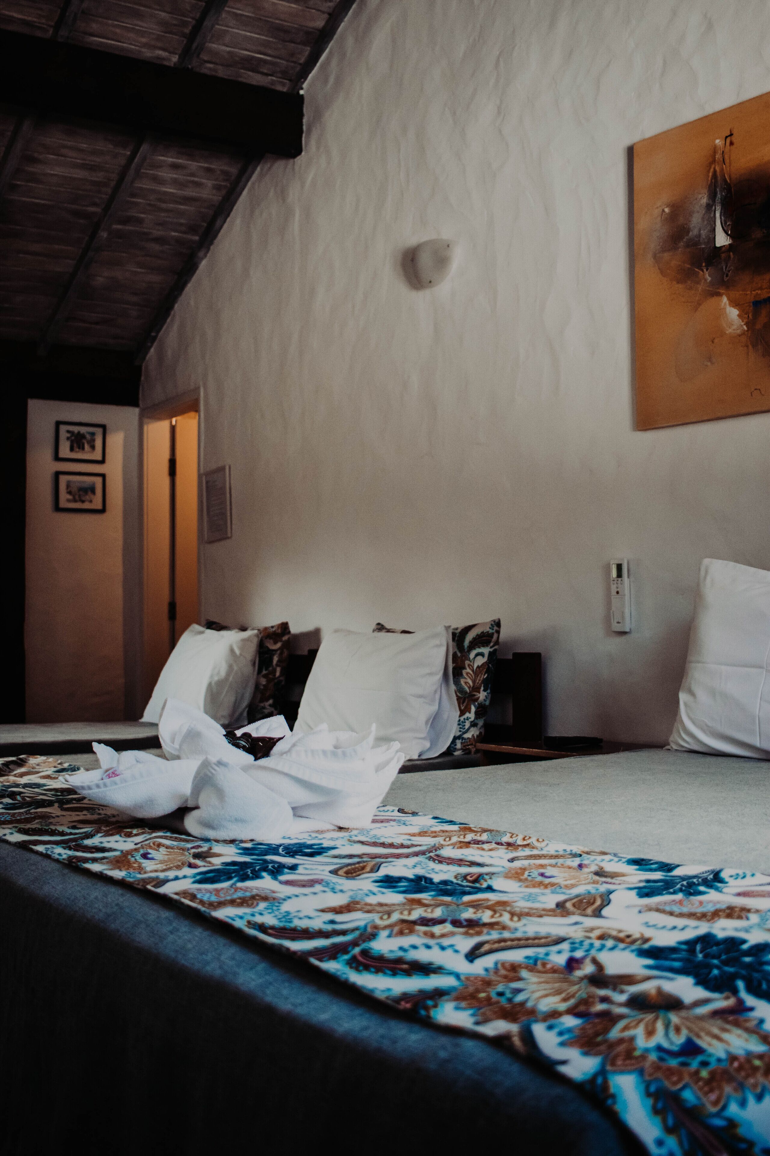 Una habitación de hotel con cama, colcha estampada, un cuadro en la pared y techo con vigas de madera.