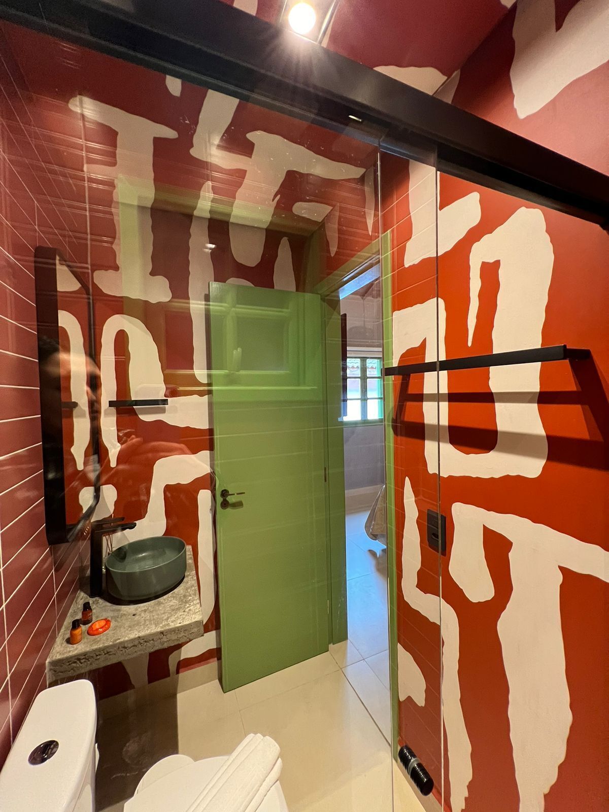 A bathroom with a green door, a white toilet, a sink with a green basin, and a mirror with a red and white abstract design.