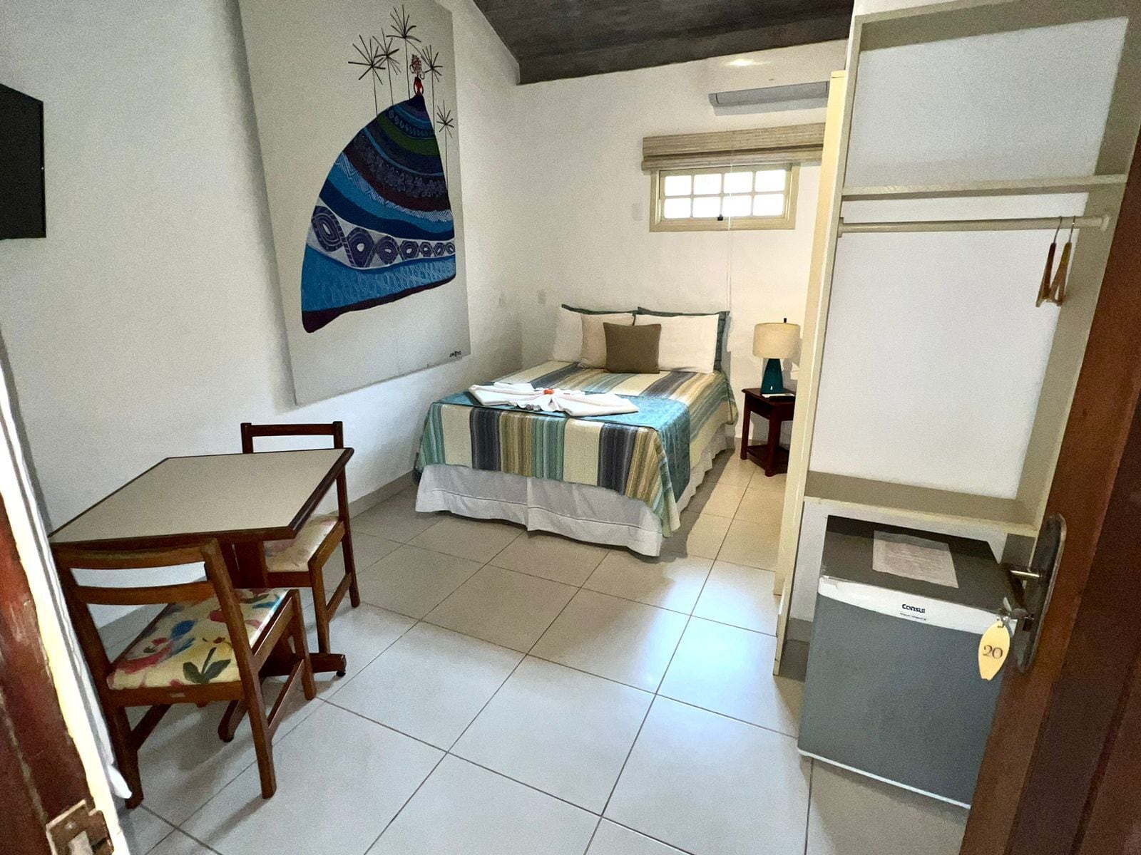 A small, well-lit room with a bed, a chair, a table, and a refrigerator.