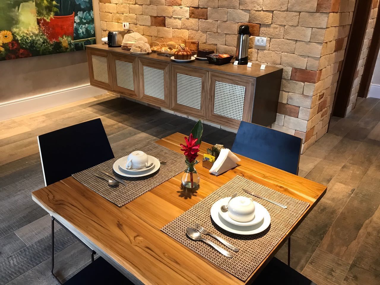 A dining area with a wooden table set for two, featuring placemats, cutlery, and a vase with flowers. A buffet counter with various food items is visible in the background.
