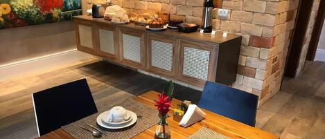 A dining area with a wooden table set for two, featuring placemats, cutlery, and a vase with flowers. A buffet counter with various food items is visible in the background.