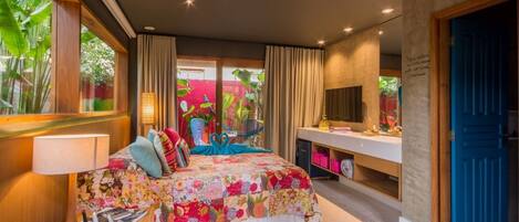 A bedroom with a large bed, a desk, a chair, and a TV. There is a window with curtains and a view of greenery outside.