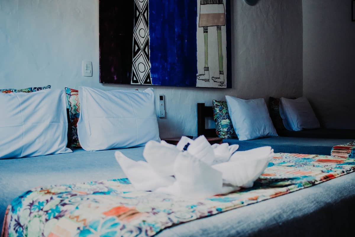 A neatly made bed with white linens and a floral-patterned blanket. There is a wall-mounted artwork above the bed and a power outlet on the wall.