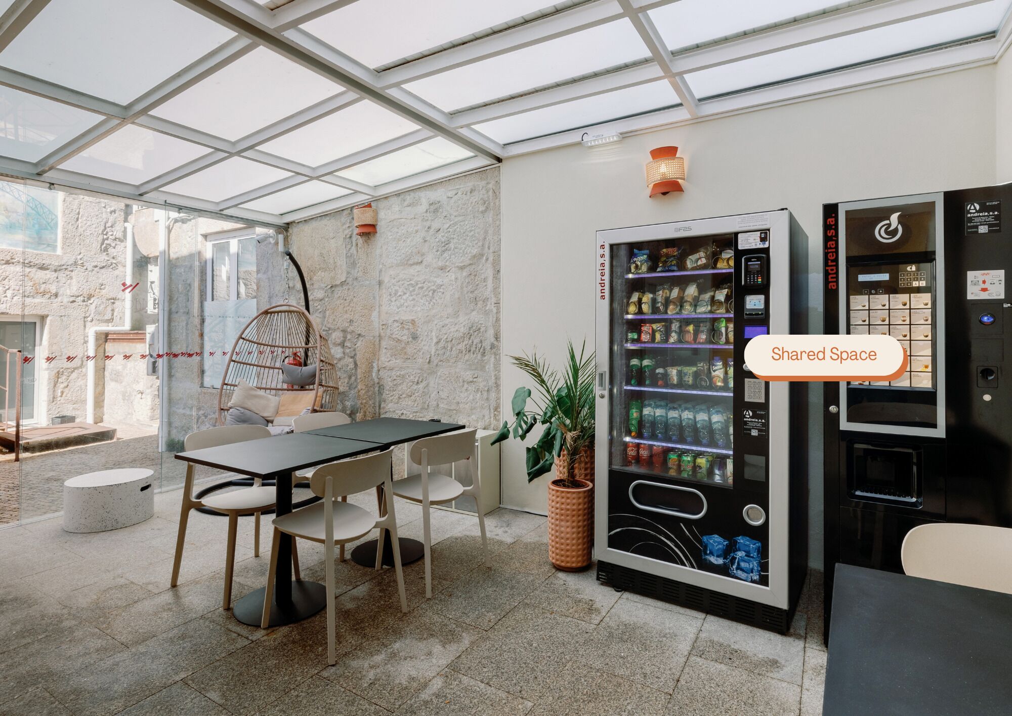 Een moderne, overdekte ruimte met glazen dak, een zithoek met tafels en stoelen, en automaten.